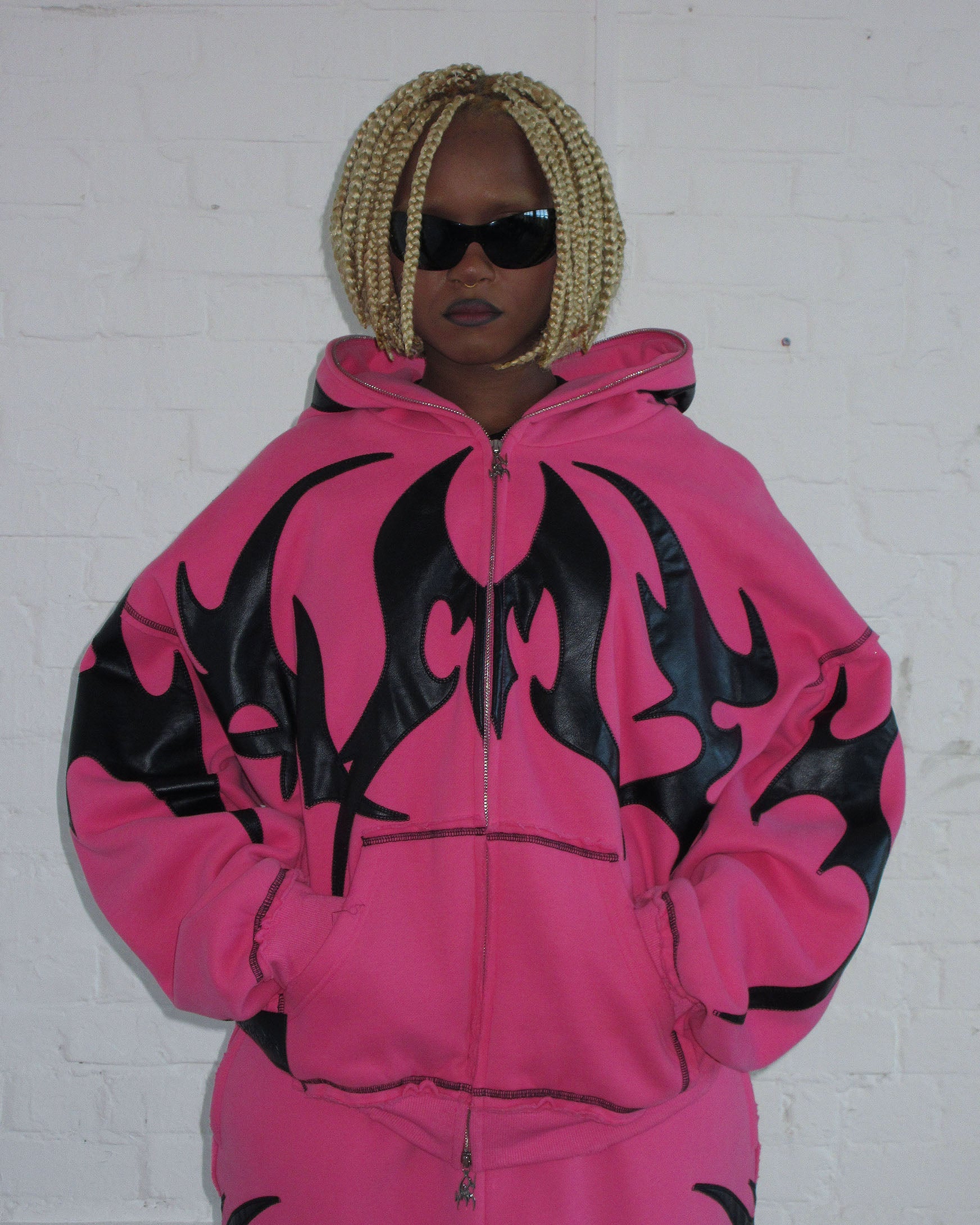 Person wears the NAMED COLLECTIVE® FLAME ZIP HOODIE PINK POISON with hands in pockets, blonde braids, and dark sunglasses.