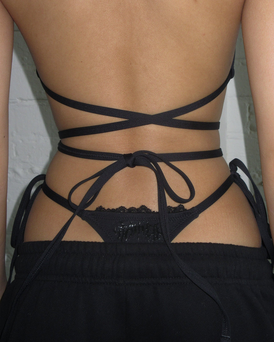 A person wears NAMED COLLECTIVE® BAD ROMANCE BIKINI BOTTOMS BLACK, shown from the back with tie details and lace trim.