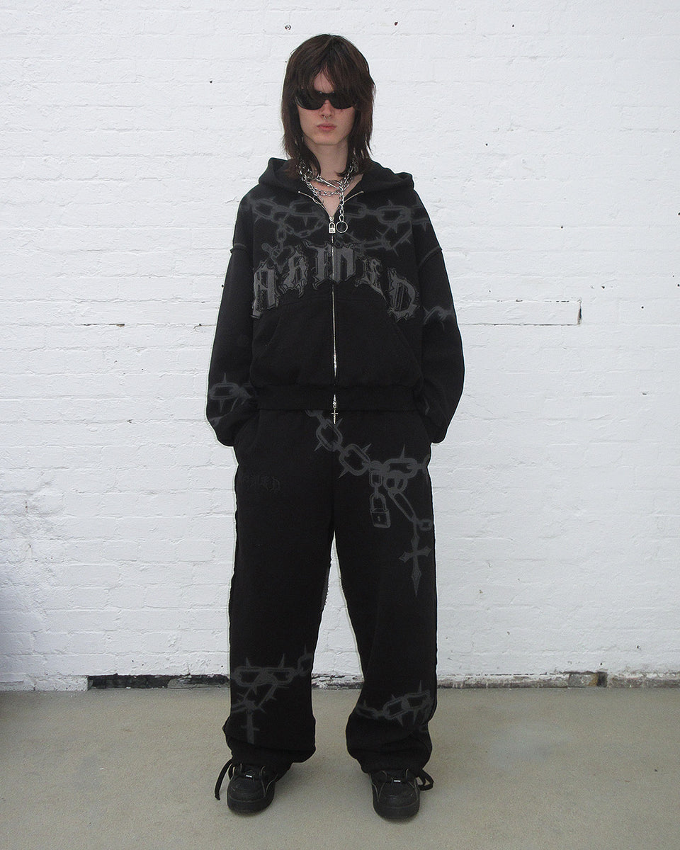 Wide shot of model wearing Chains oversized sweatpants black standing in front of a white brick wall. 