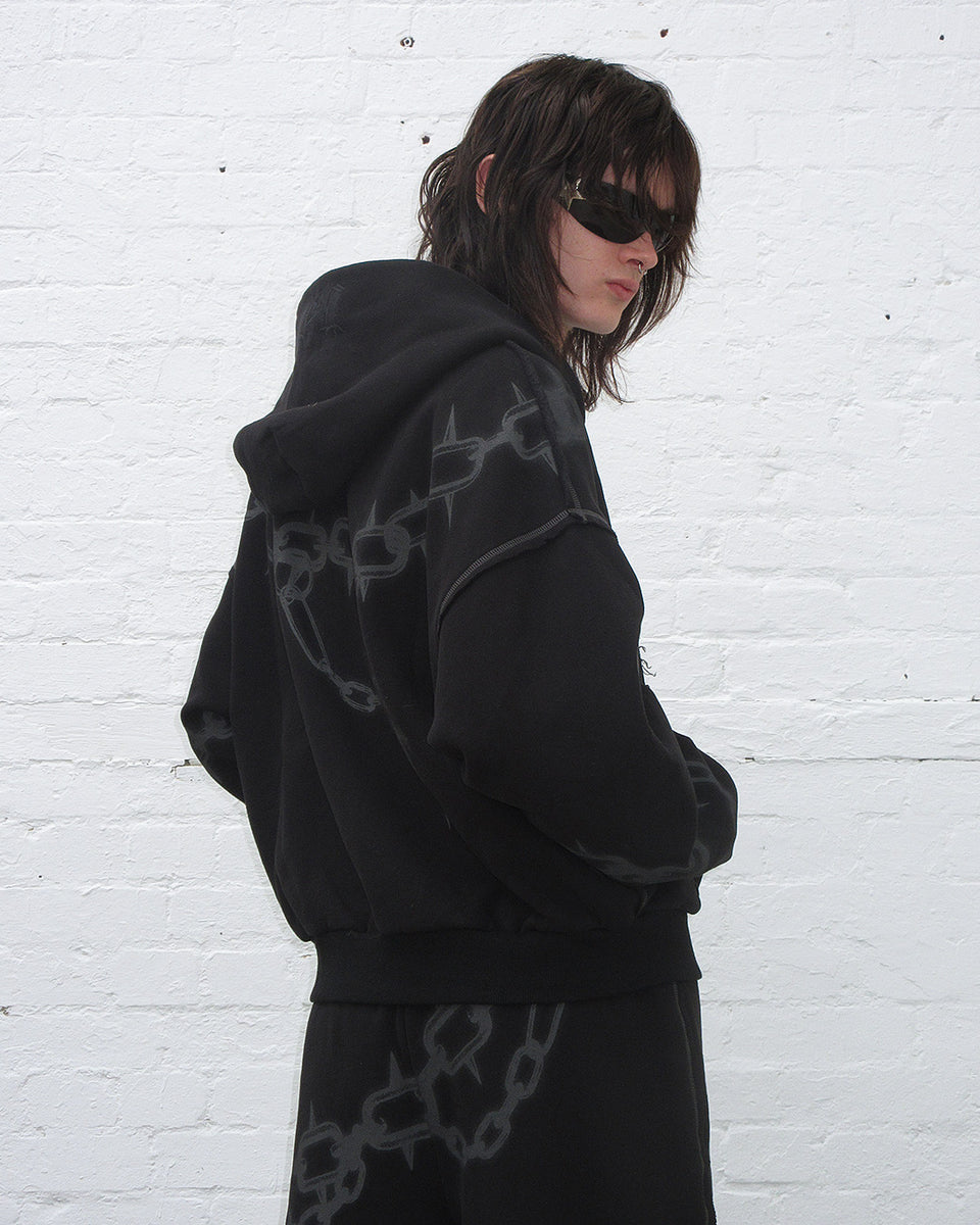 Model wearing Chains zip hoodie black , styled with sunglasses against a white brick wall.
