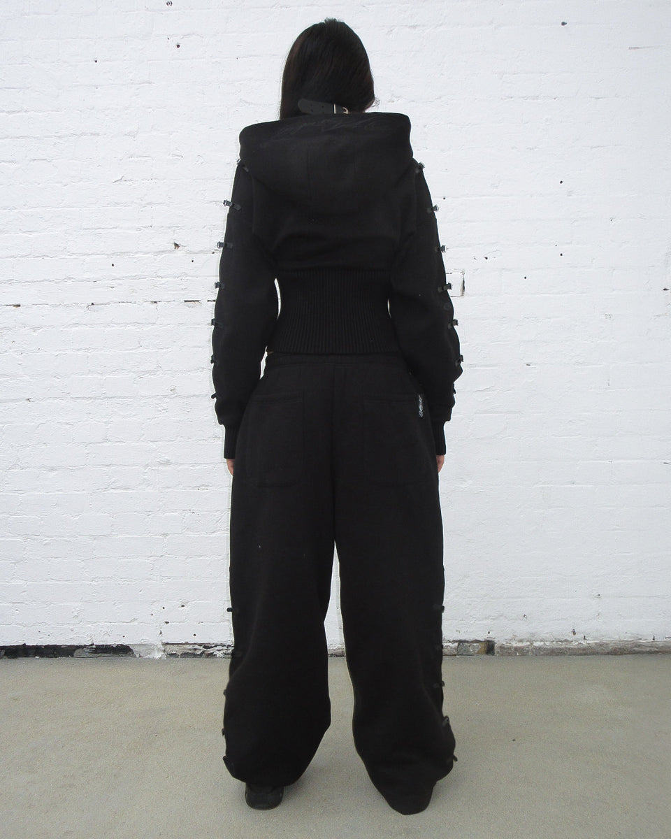 Back view of a model wearing Crush buckle fitted waist zip hoodie black with stylish pants.