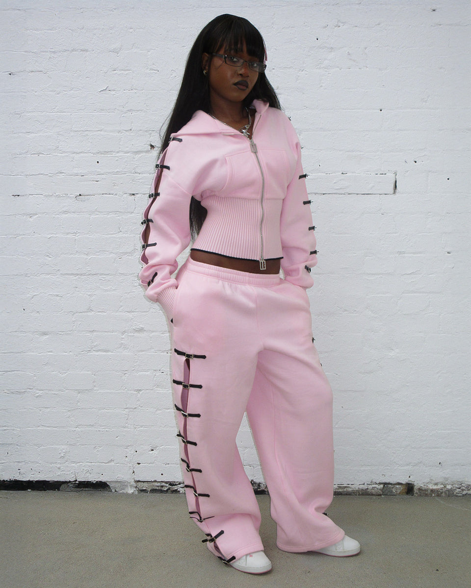 Model wearing crush buckle oversized sweatpants baby pink with matching hoodie in front of a white wall.