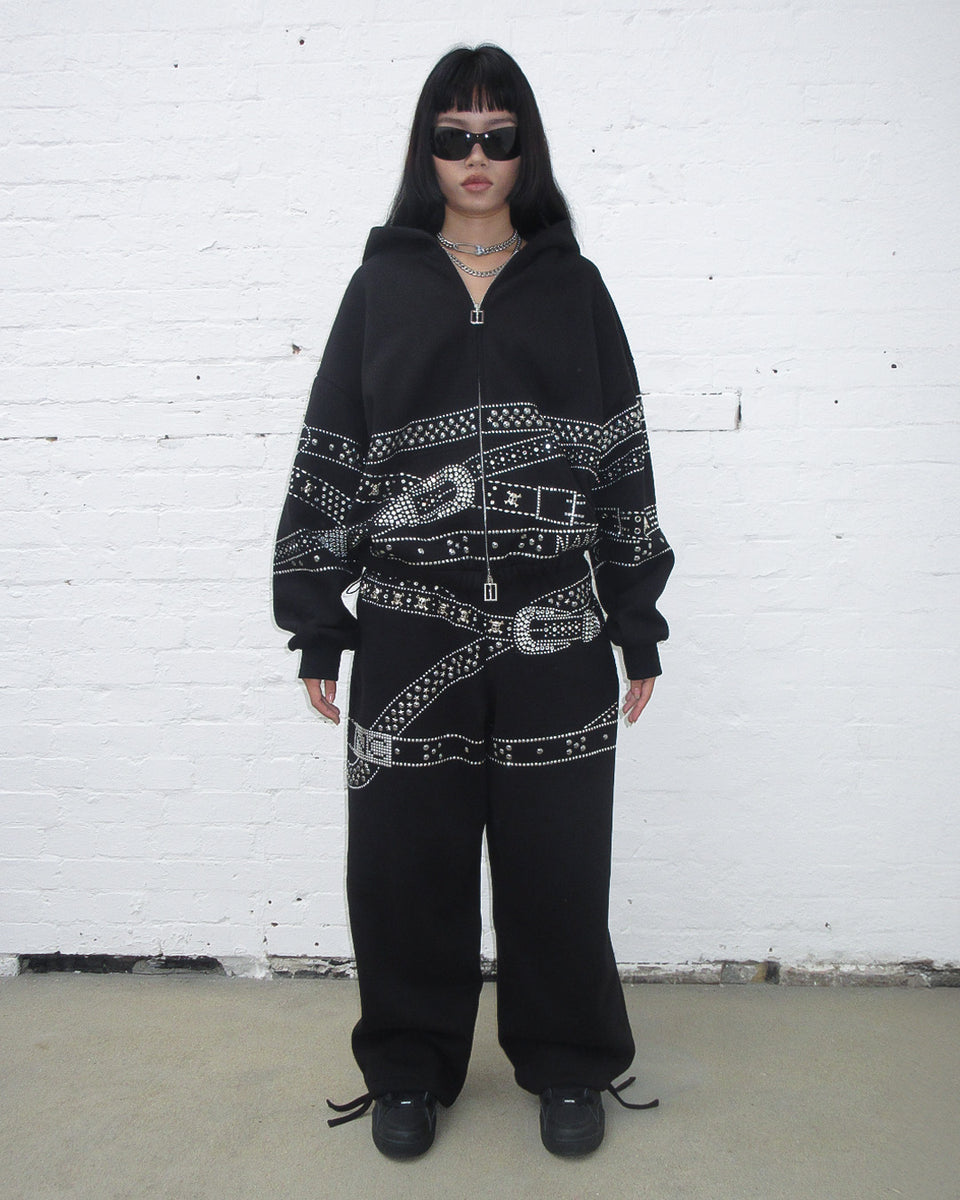 Crush studded belt oversized sweatpants black worn by model with black sunglasses against a white brick wall.