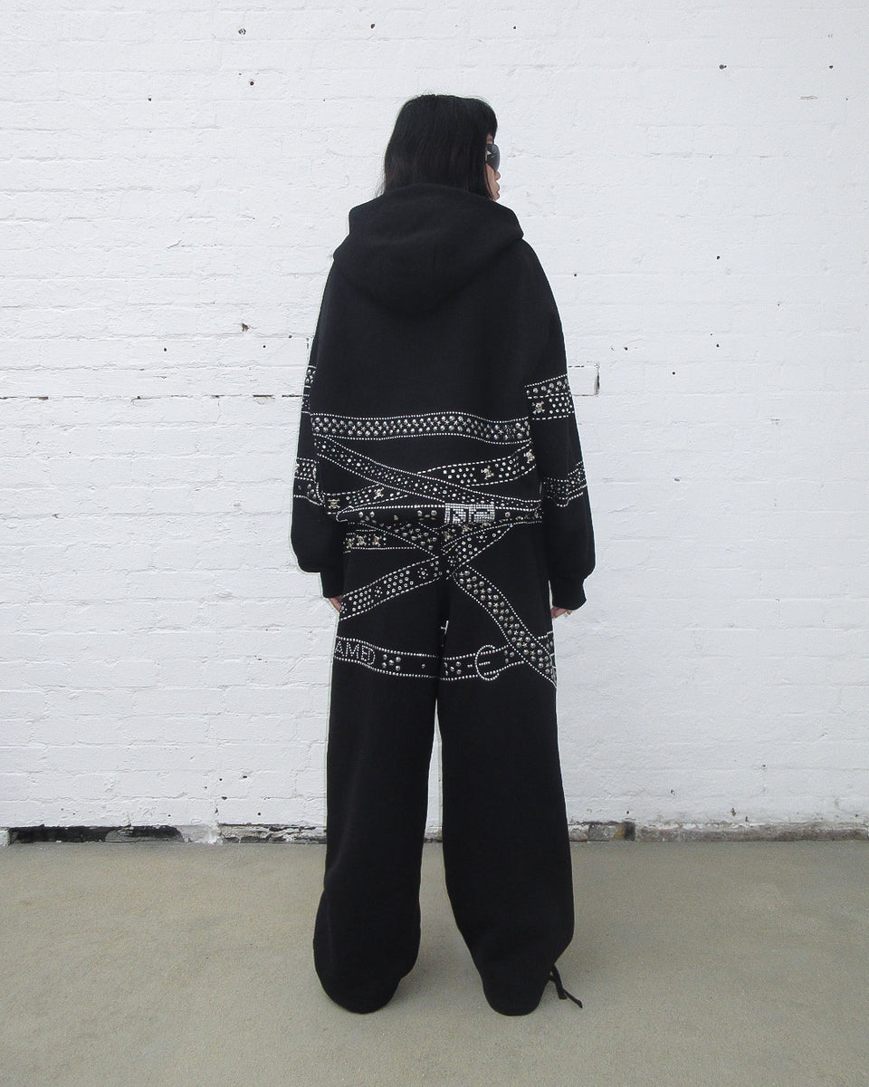 Crush studded belt zip hoodie black worn from the back against a white brick wall.