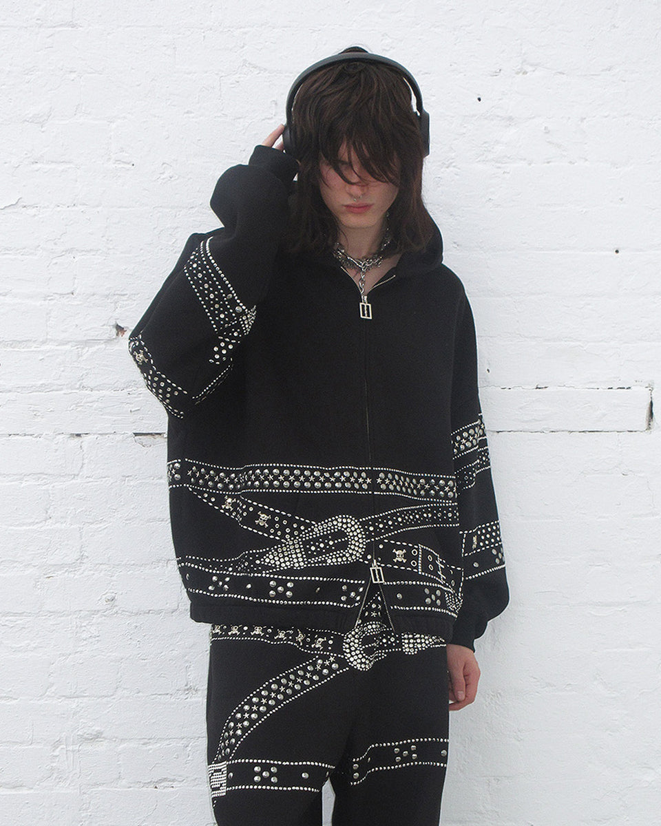 Crush studded belt zip hoodie black worn by model with headphones against a white brick background.