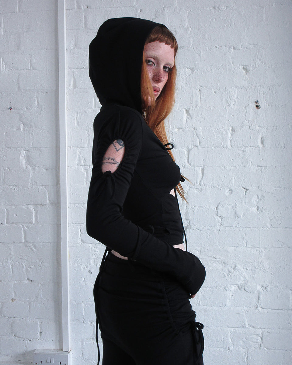 A person with long red hair wears the NAMED COLLECTIVE® ENVY SCULPT STRETCH TIE HOODIE BLACK, standing by a white brick wall.