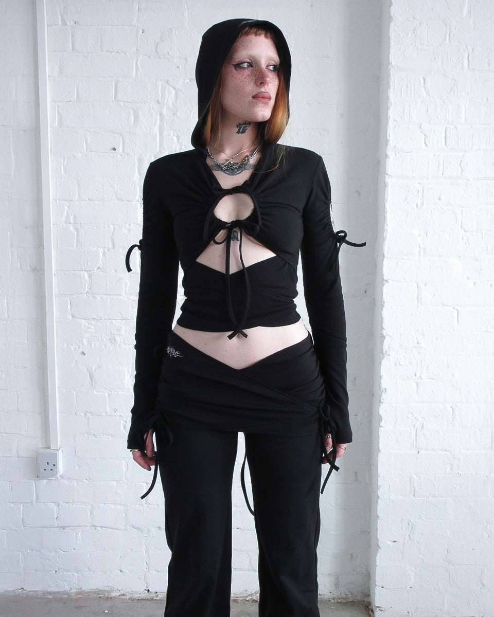 Model wearing the ENVY SCULPT STRETCH TIE HOODIE BLACK by NAMED COLLECTIVE® against a white brick wall.
