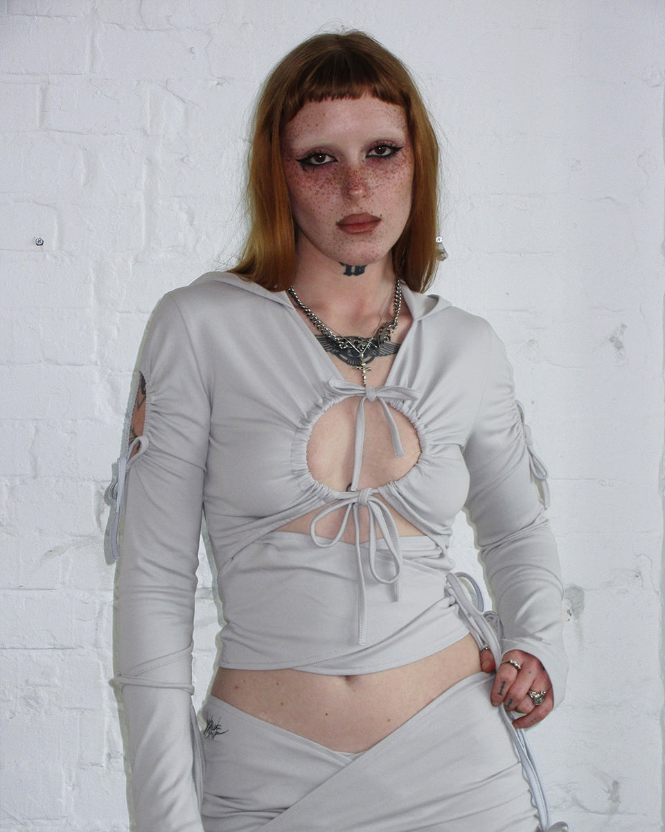 Person with red hair and freckles wears NAMED COLLECTIVE® ENVY SCULPT STRETCH TIE HOODIE in mist grey against a white wall.