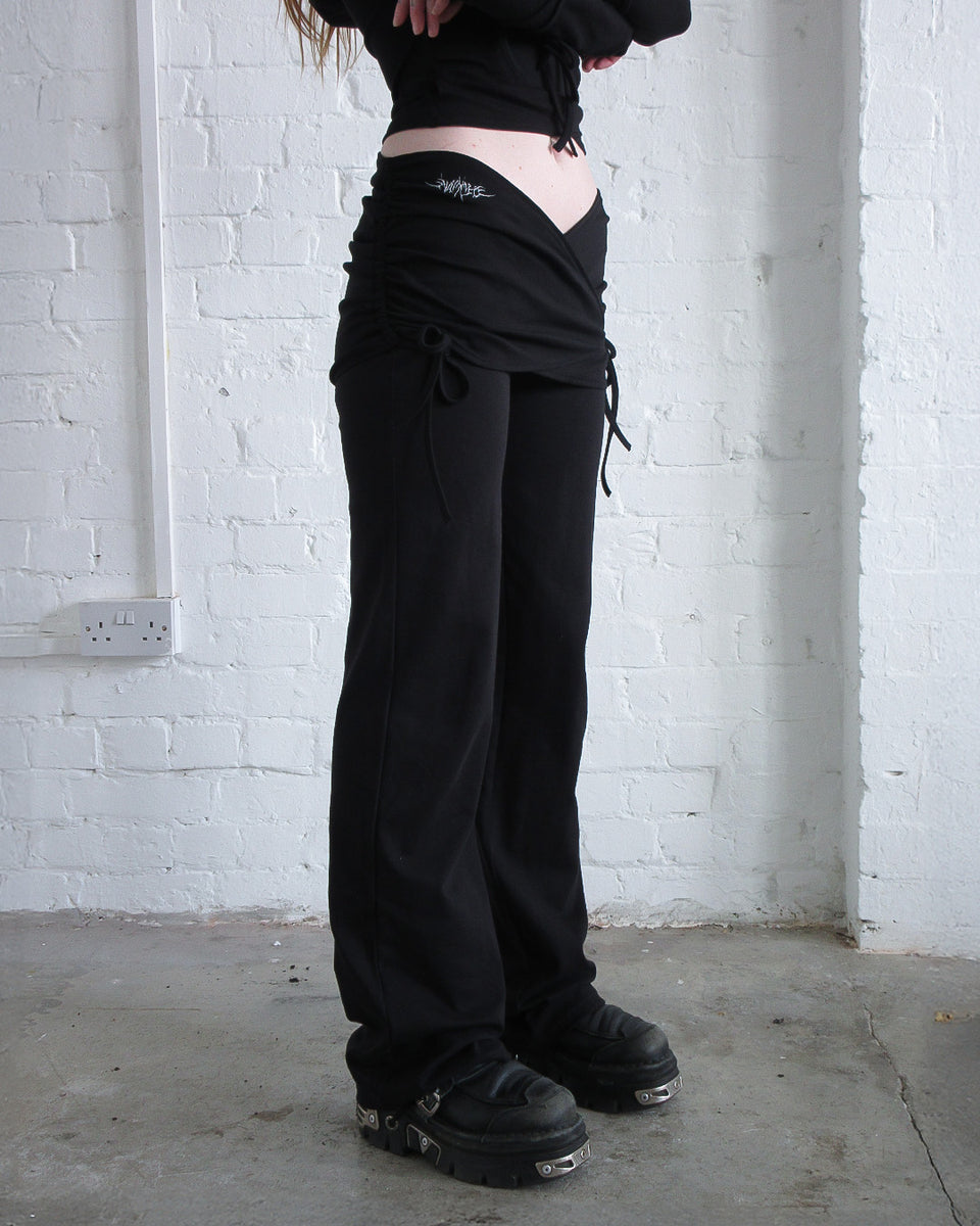 Model wears the ENVY SCULPT STRETCH TWO PIECE SKIRT TROUSER SET BLACK by NAMED COLLECTIVE® against a white brick wall.
