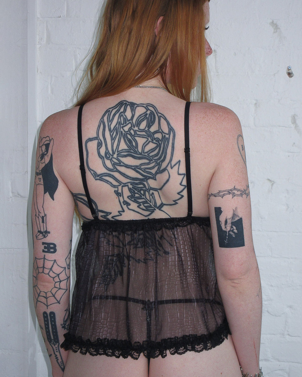 Wearing NAMED COLLECTIVE® ENVY Sheer Snakeskin Mesh Cami Top Charcoal, seen from the back against a white brick wall.