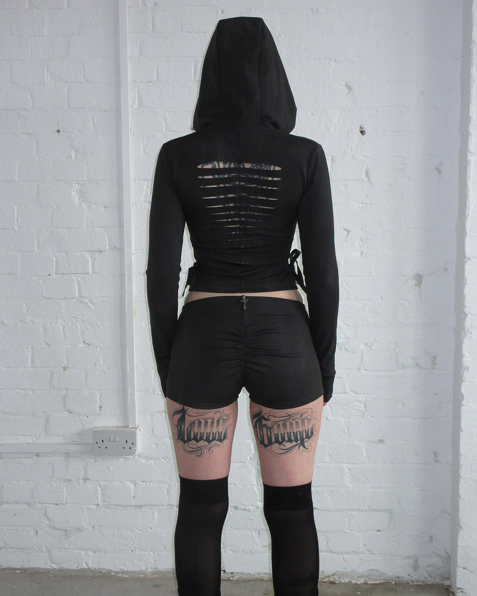 Person in ENVY SNAKESKIN HOTPANT SHORTS CHARCOAL by NAMED COLLECTIVE® stands against a white brick wall.
