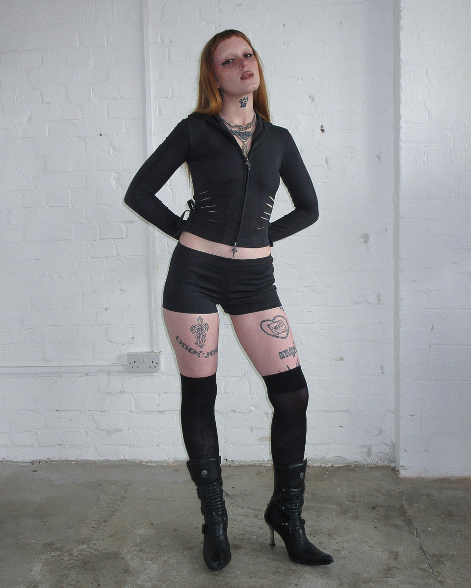 Model with visible tattoos wears NAMED COLLECTIVE® ENVY SNAKESKIN HOTPANT SHORTS CHARCOAL, styled with a black outfit and heels.