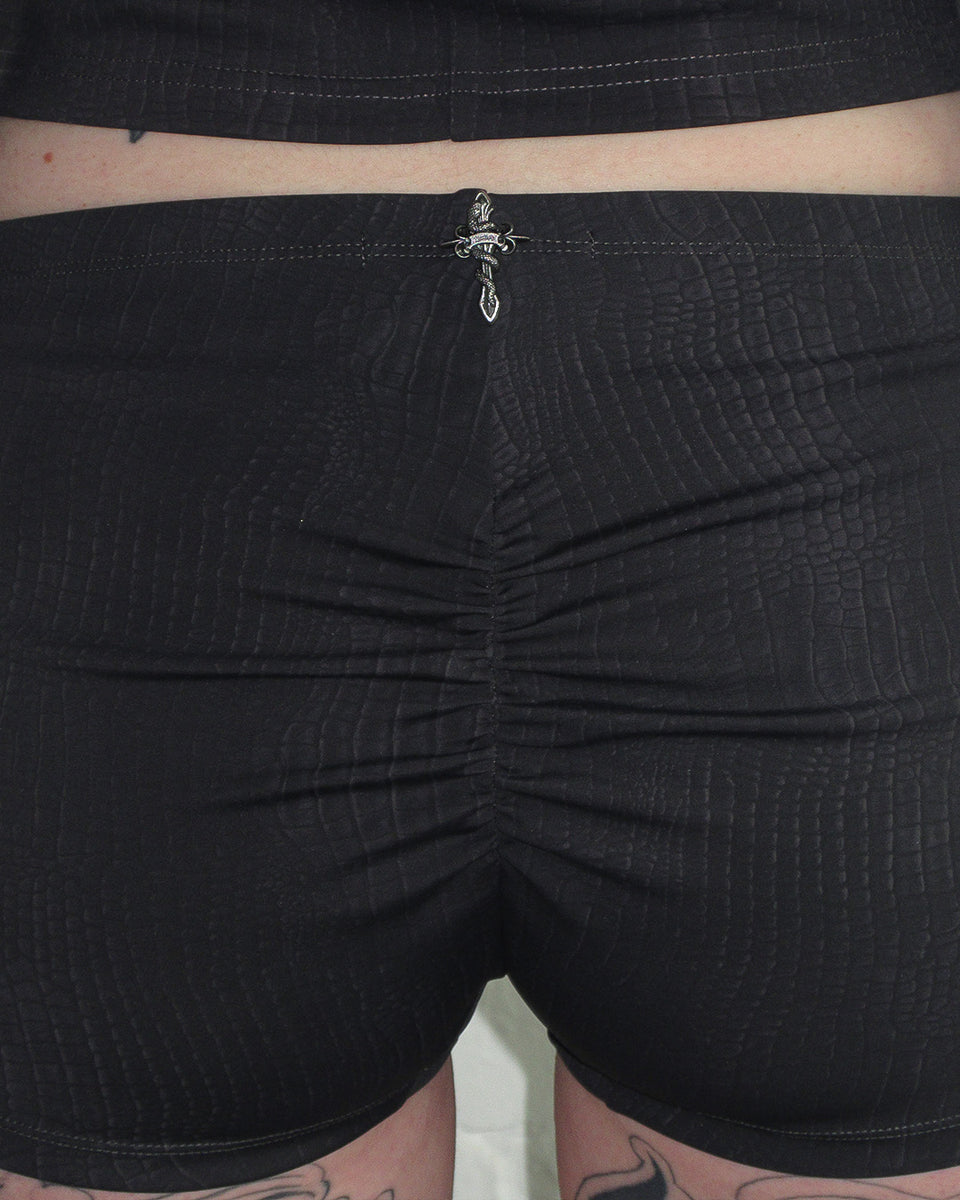 Close-up of a person wearing NAMED COLLECTIVE® ENVY SNAKESKIN HOTPANT SHORTS CHARCOAL with a silver sword pin at the waist.