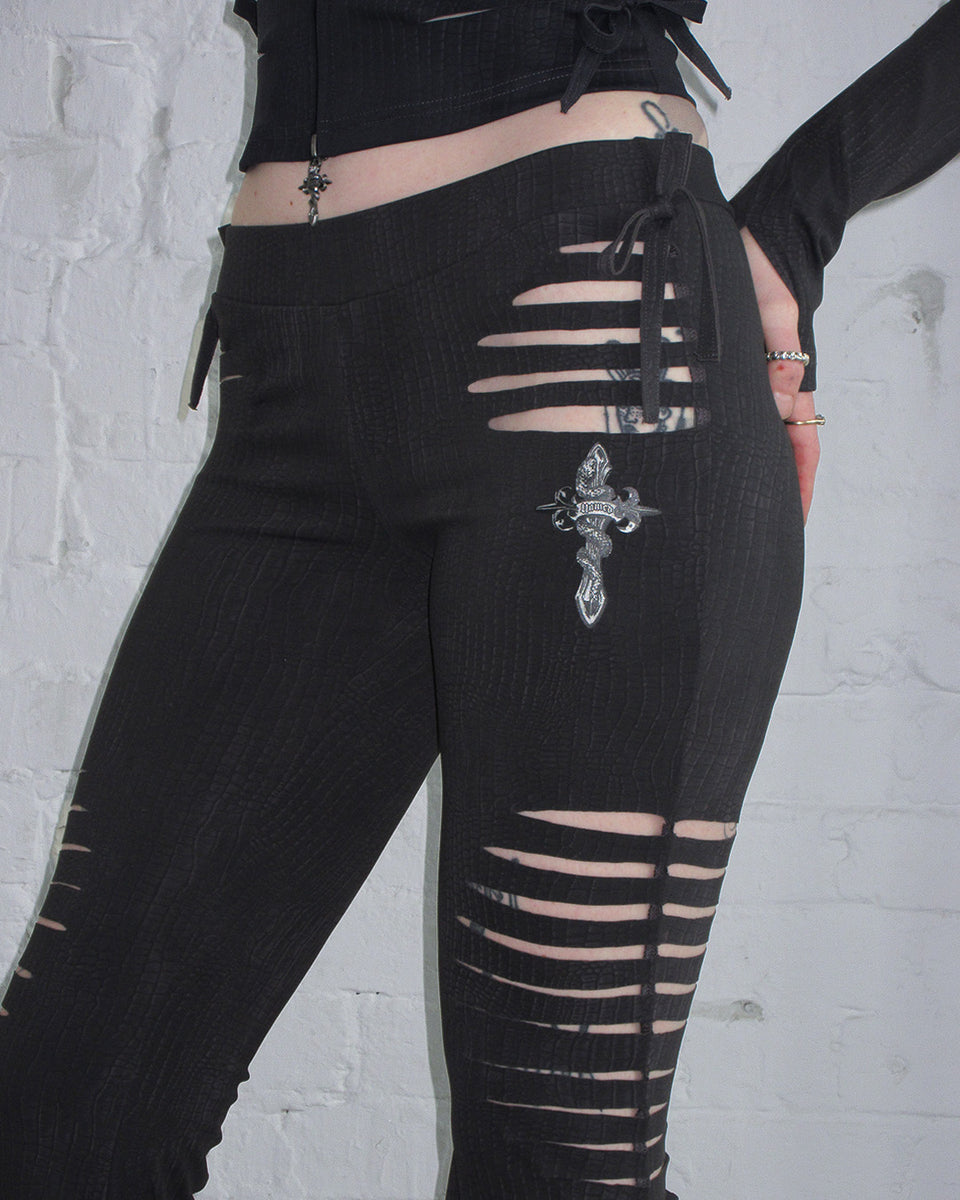 Person wears NAMED COLLECTIVE® ENVY SNAKESKIN SLASHED SCULPT STRETCH PANTS CHARCOAL against a white brick wall.