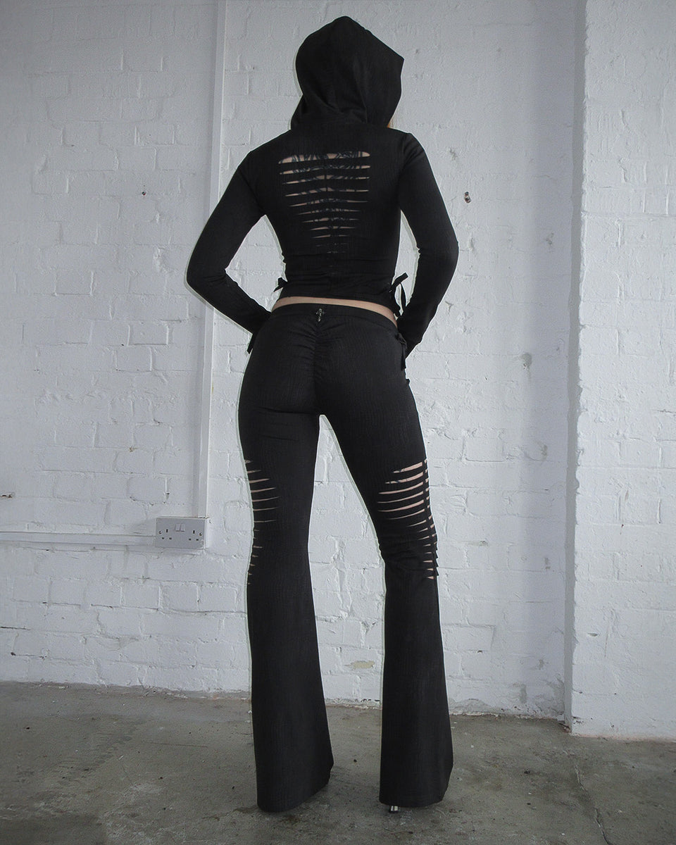 Model faces away in NAMED COLLECTIVE® ENVY SNAKESKIN SLASHED SCULPT STRETCH PANTS CHARCOAL, in a minimalist room.