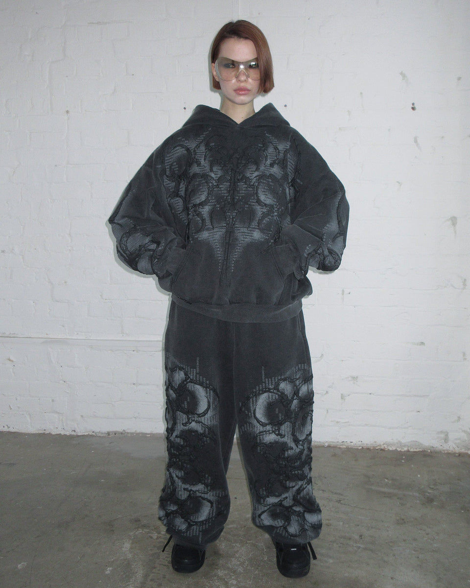 Person standing against a white brick wall wearing NAMED COLLECTIVE® EUPHORIA Puff Print Oversized Sweatpants in Washed Charcoal.