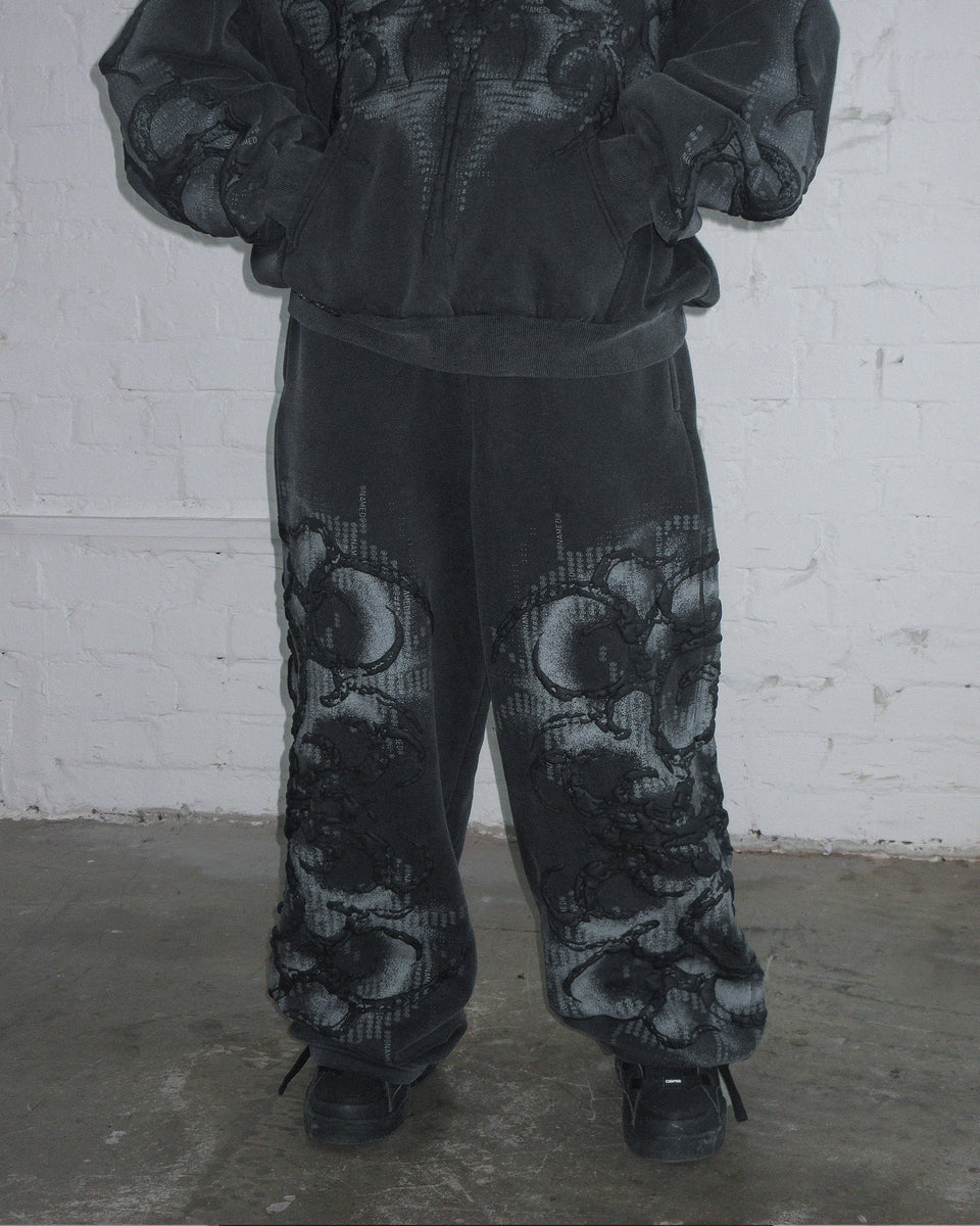 Model in NAMED COLLECTIVE® EUPHORIA PUFF PRINT OVERSIZED SWEATPANTS WASHED CHARCOAL stands by a white brick wall.