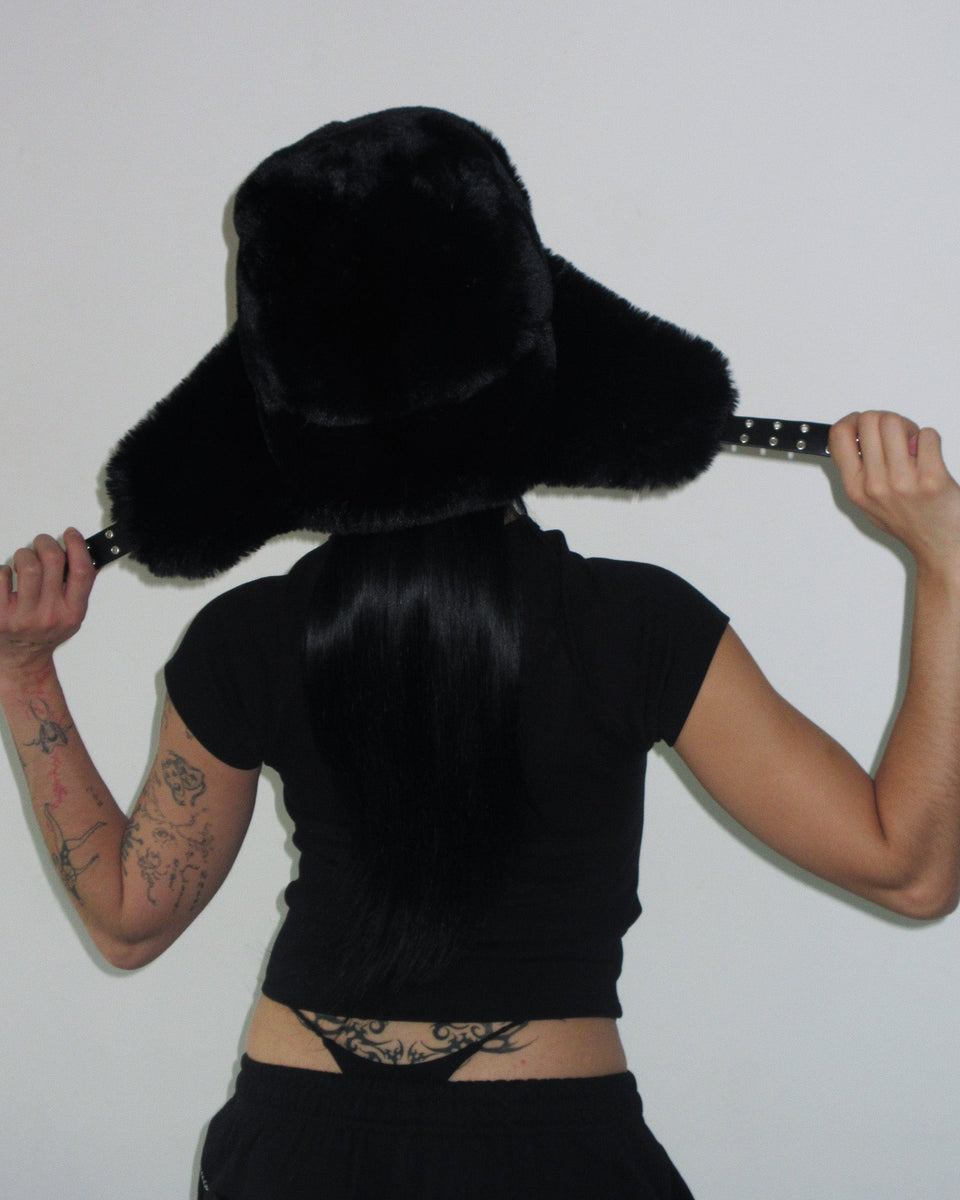 Tattooed person in a black FERAL STUDDED BABY TEE by NAMED COLLECTIVE®, black fur hat, facing away, holding hat straps.