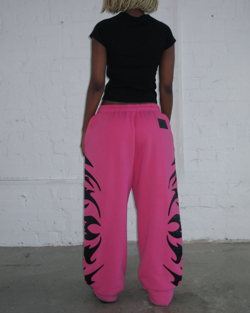 A person stands against a white wall in a black shirt and NAMED collective FLAME SWEATPANTS PINK POISON.