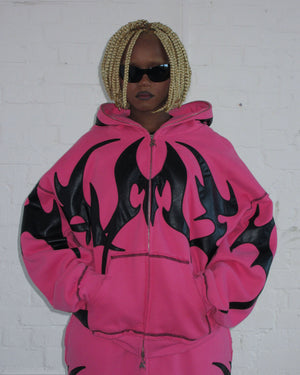 Person wears the NAMED COLLECTIVE® FLAME ZIP HOODIE PINK POISON with hands in pockets, blonde braids, and dark sunglasses.