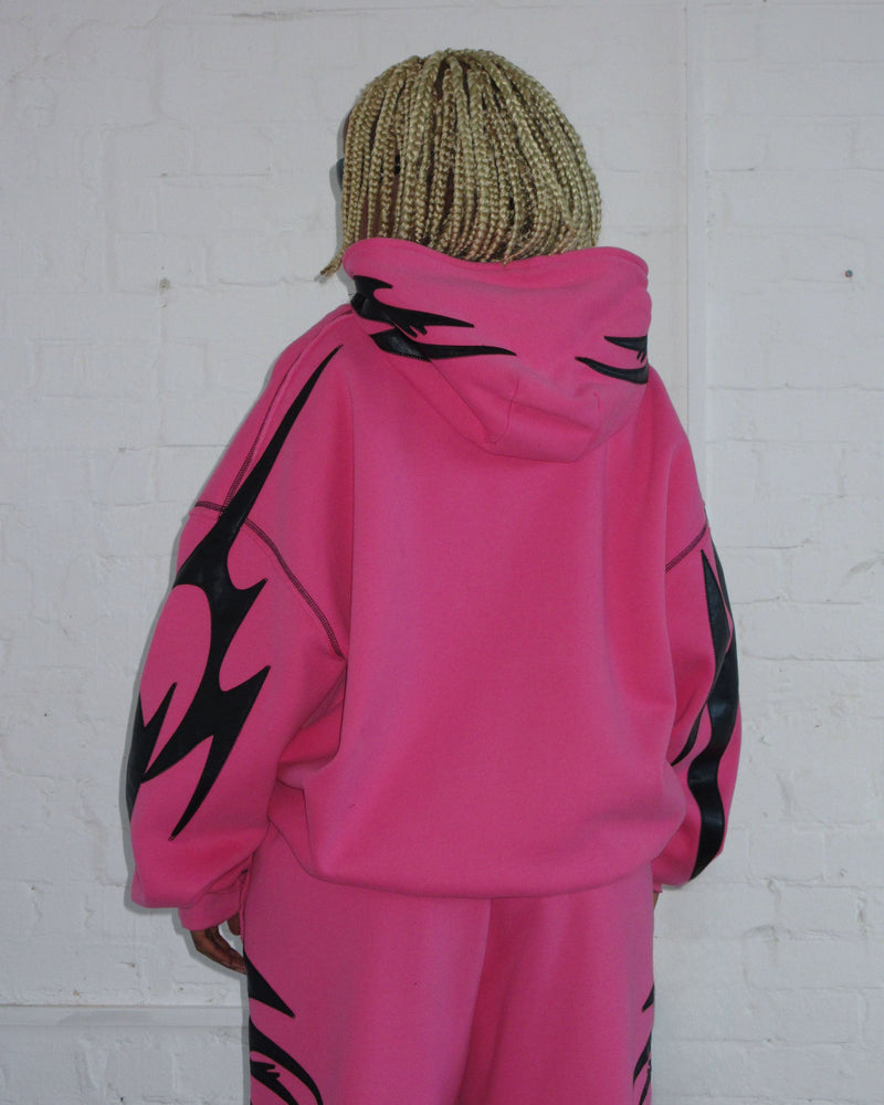 Person with blonde braids wearing NAMED COLLECTIVE® FLAME ZIP HOODIE PINK POISON, facing a white wall.