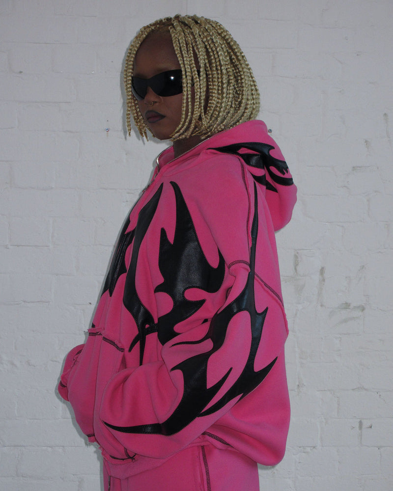 Person wears the NAMED COLLECTIVE® FLAME ZIP HOODIE PINK POISON, sunglasses, and blonde box braids by a white brick wall.