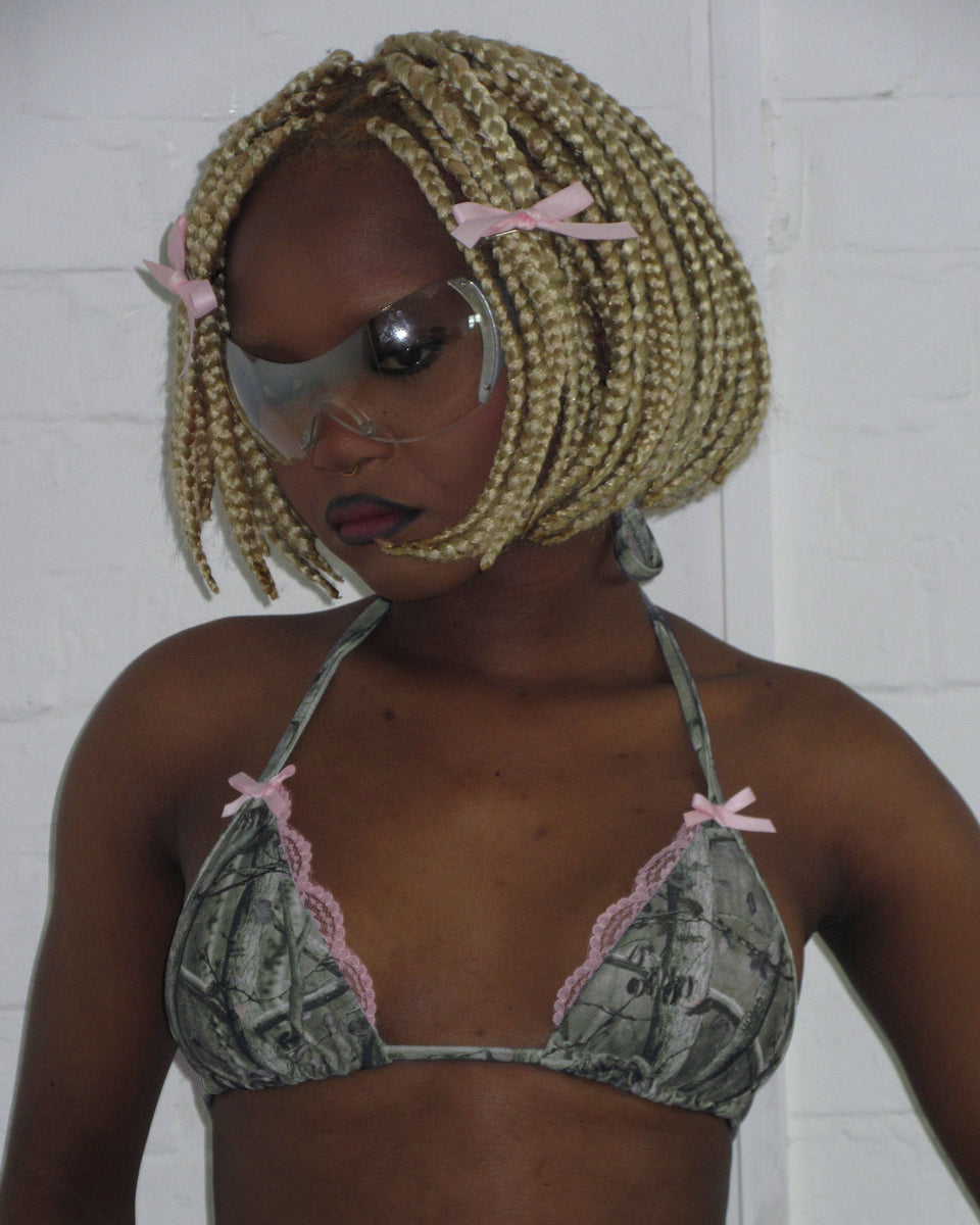 Person with blonde braided bob, large clear glasses, wears NAMED COLLECTIVE® FORBIDDEN BIKINI TOP NATURAL, against white wall.