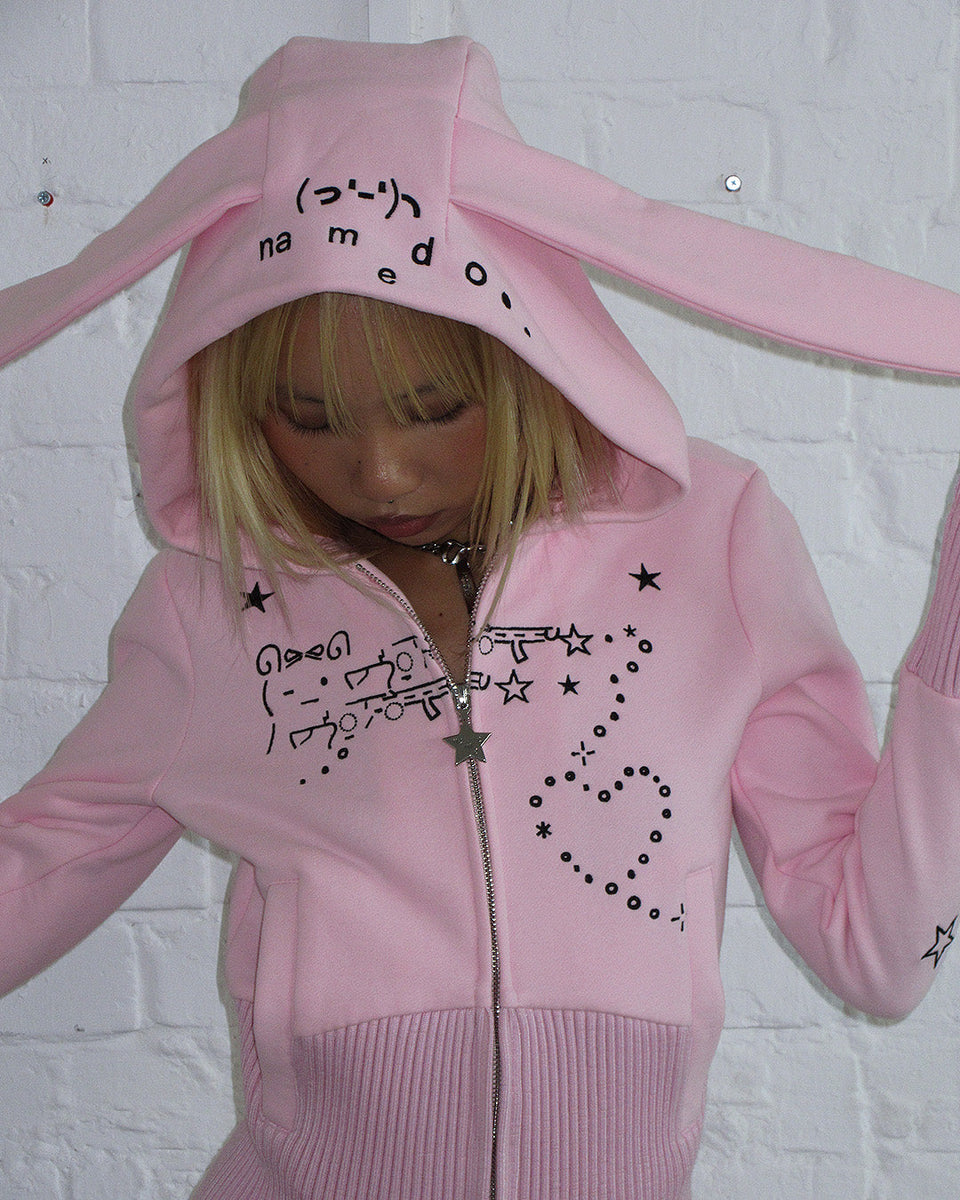 Model wears NAMED COLLECTIVE® MALFUNCTION BUNNY FITTED HOODIE BABY PINK (DETACHABLE EARS) by a white brick wall.