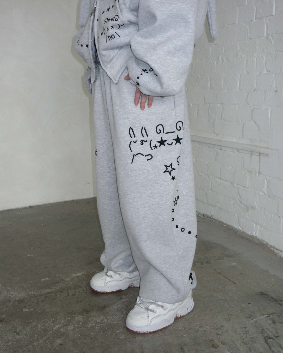 Person wearing NAMED COLLECTIVE® MALFUNCTION SWEATPANTS GREY MARL and white sneakers, standing by a white brick wall.