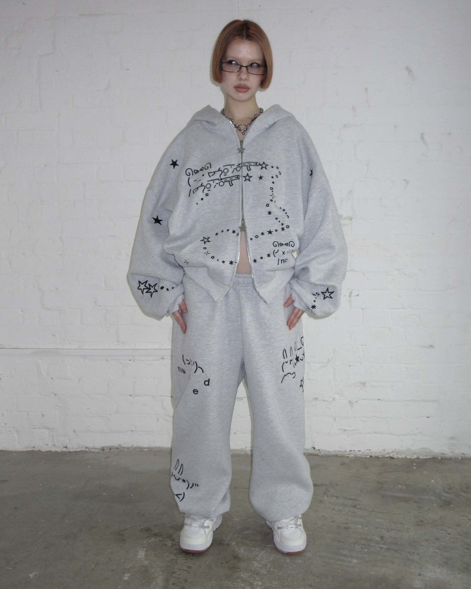 Person stands against a white brick wall wearing NAMED COLLECTIVE® MALFUNCTION SWEATPANTS GREY MARL and white sneakers.