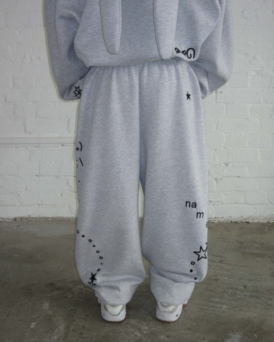 Model wears NAMED COLLECTIVE® MALFUNCTION SWEATPANTS GREY MARL, standing by a white brick wall and concrete floor.
