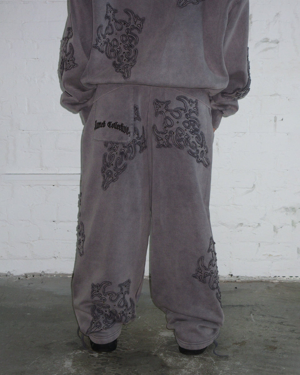 Model wears NAMED COLLECTIVE® SINNER STUDDED OVERSIZED SWEATPANTS in grey vintage wash against a white brick wall.