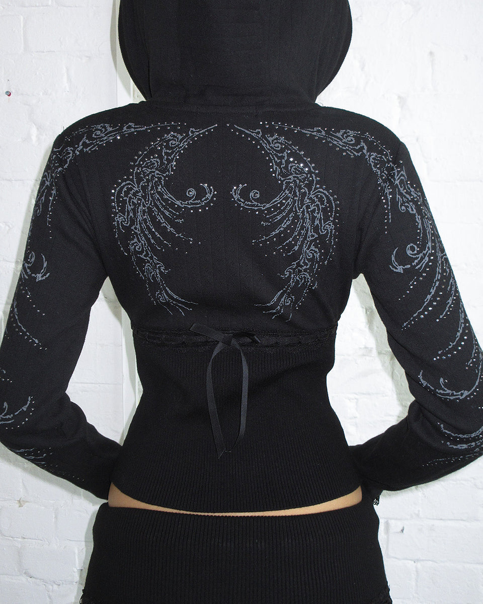 Person wears the NAMED COLLECTIVE® WHISPER RIBBON POINTELLE SCULPT HOODIE BLACK, facing a white brick wall.