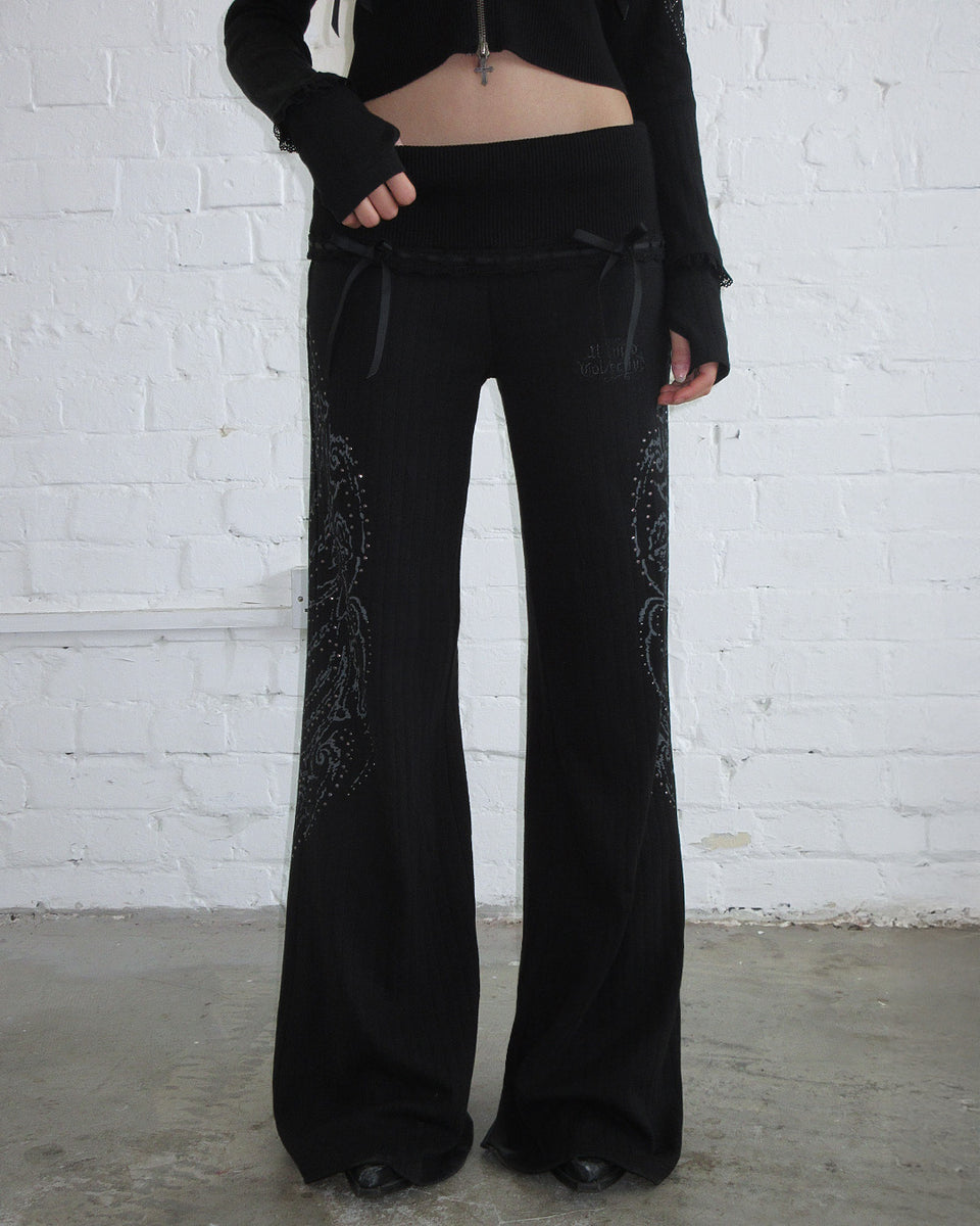 Person in black WHISPER RIBBON POINTELLE SCULPT PANTS by NAMED COLLECTIVE®, standing by a white brick wall.