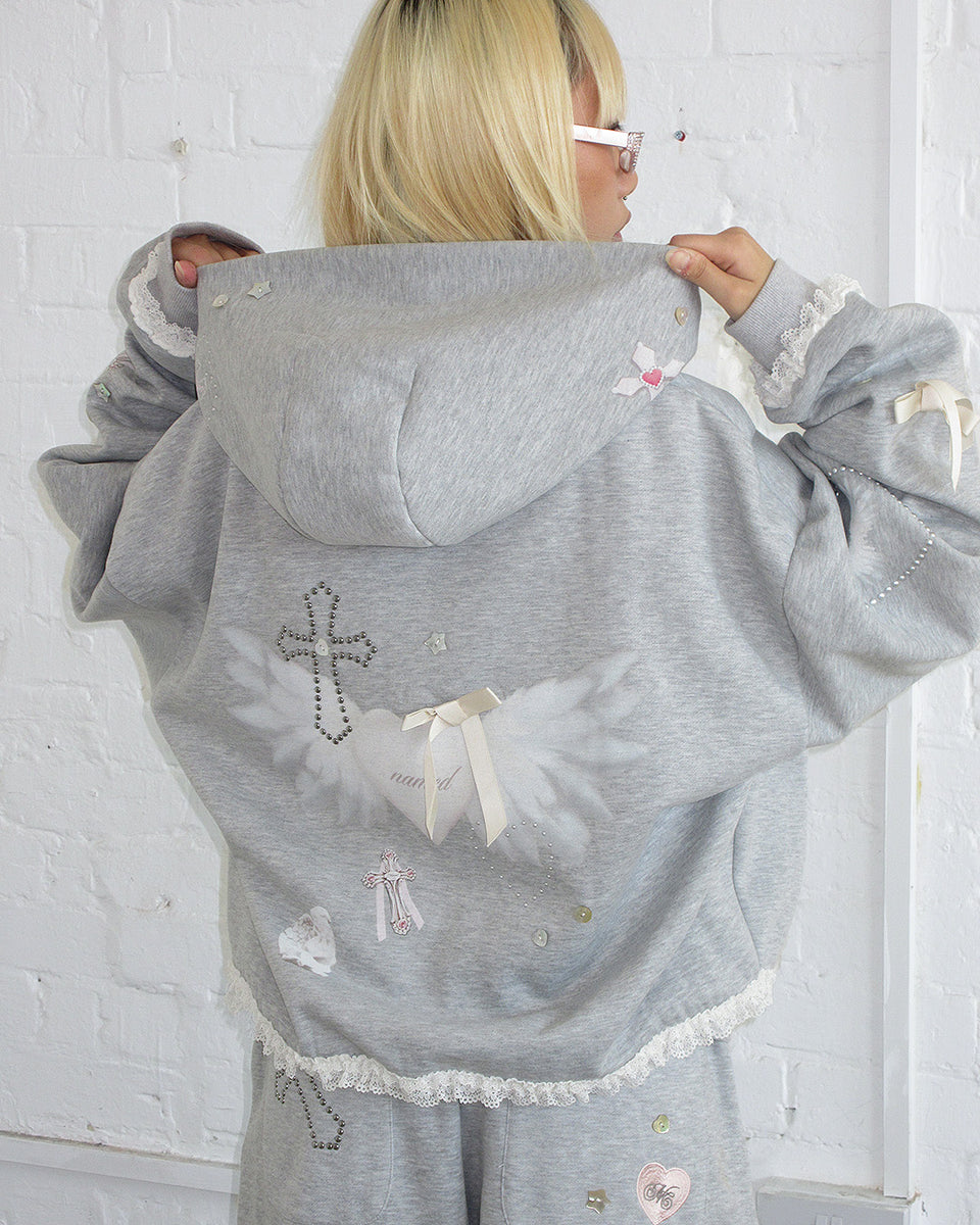 Person wearing the NAMED COLLECTIVE® WHISPER ZIP HOODIE GREY MARL with angel wings and cross design against a white brick wall.